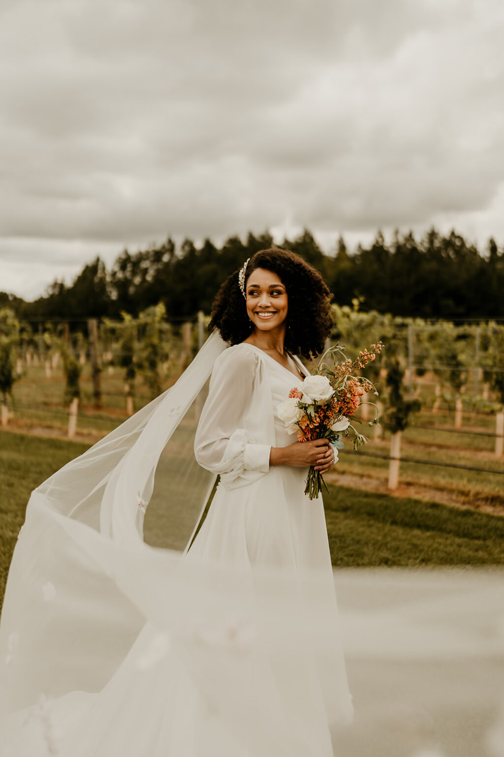 A woman in white dress holding flowers near trees.