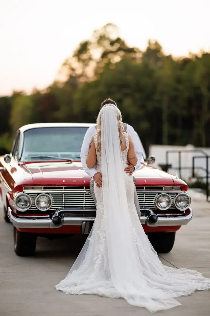 A bride in a white dress standing next to a red car.