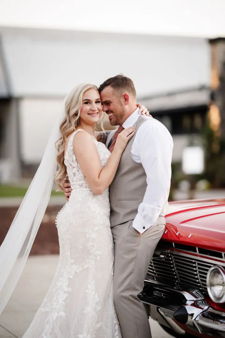 A bride and groom pose for the camera in front of a red car.