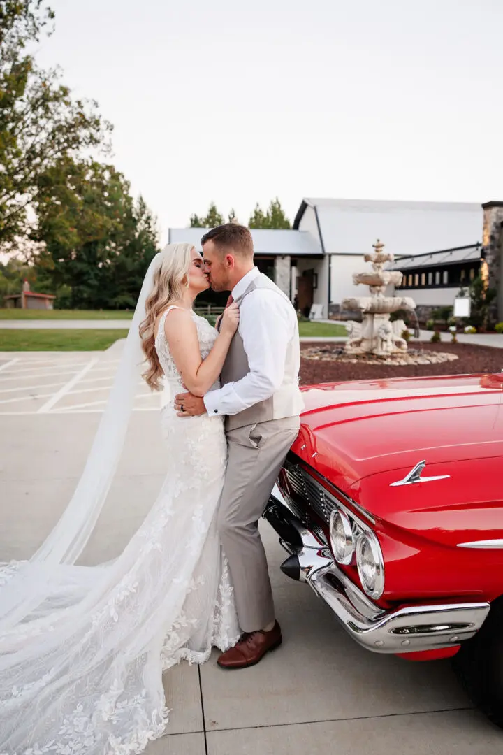 A bride and groom kissing in front of a red car.