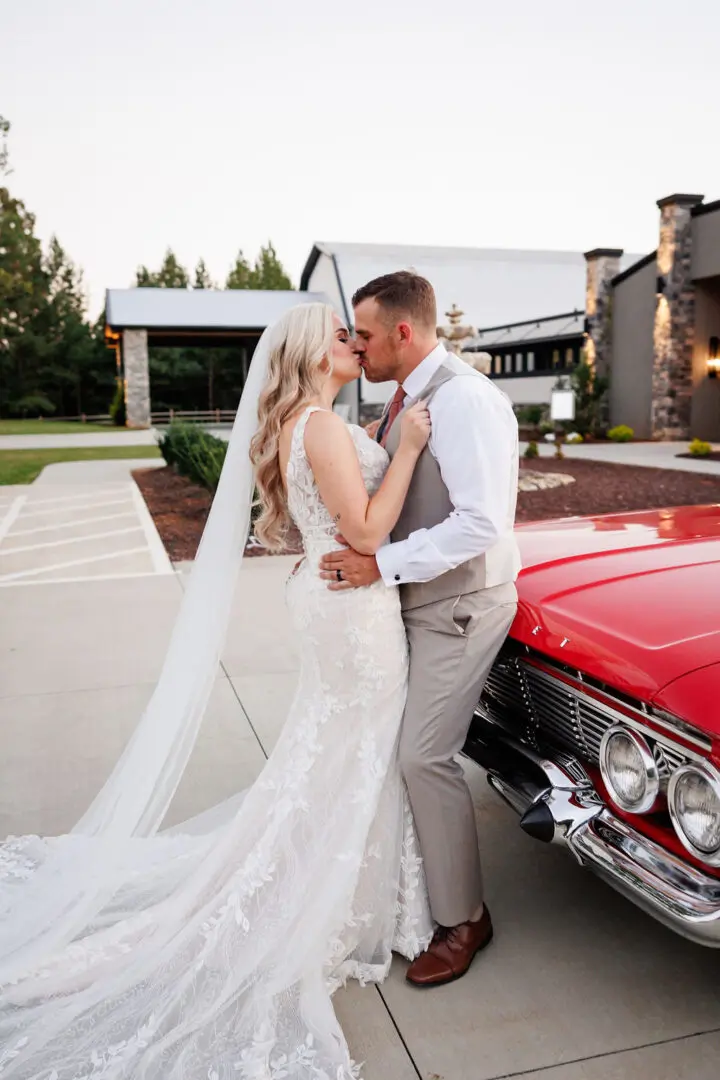 A bride and groom kissing in front of a red car.