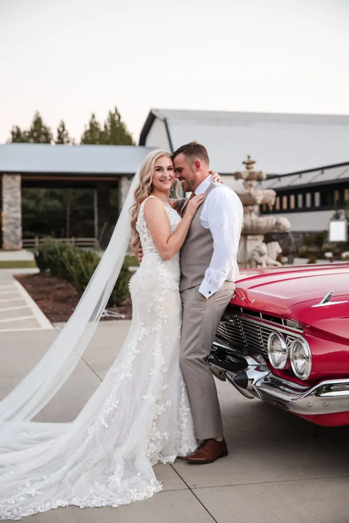 A bride and groom pose in front of an old red car.