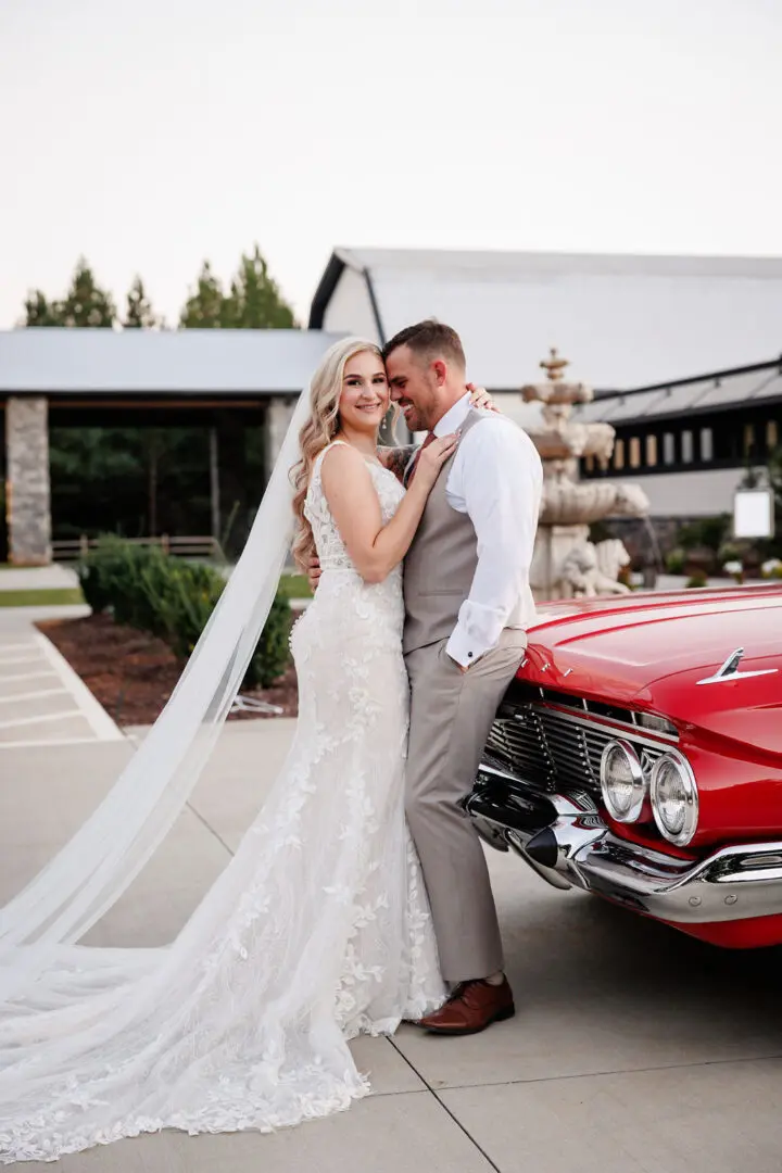 A bride and groom pose for a picture in front of an old red car.