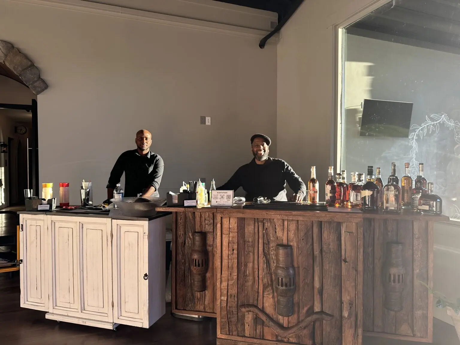 Two men standing behind a bar with bottles of alcohol.
